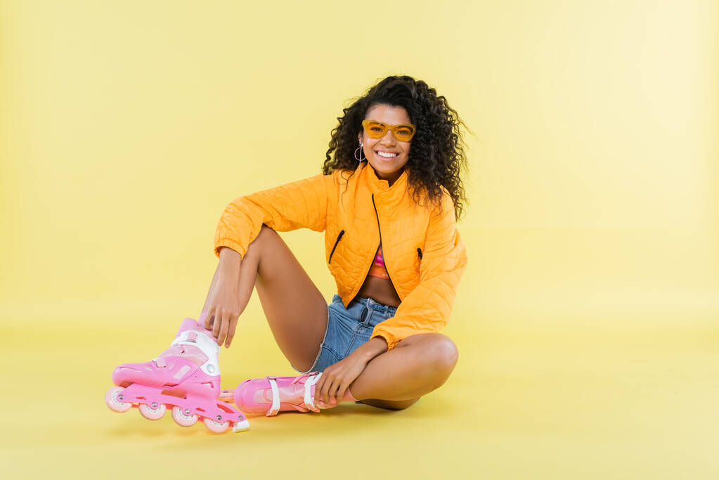 full length of cheerful african american young woman in pink roller skates sitting on yellow - Photo, Image