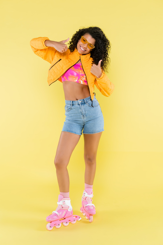 full length of cheerful african american young woman in pink roller skates showing thumbs up on yellow - Photo, Image