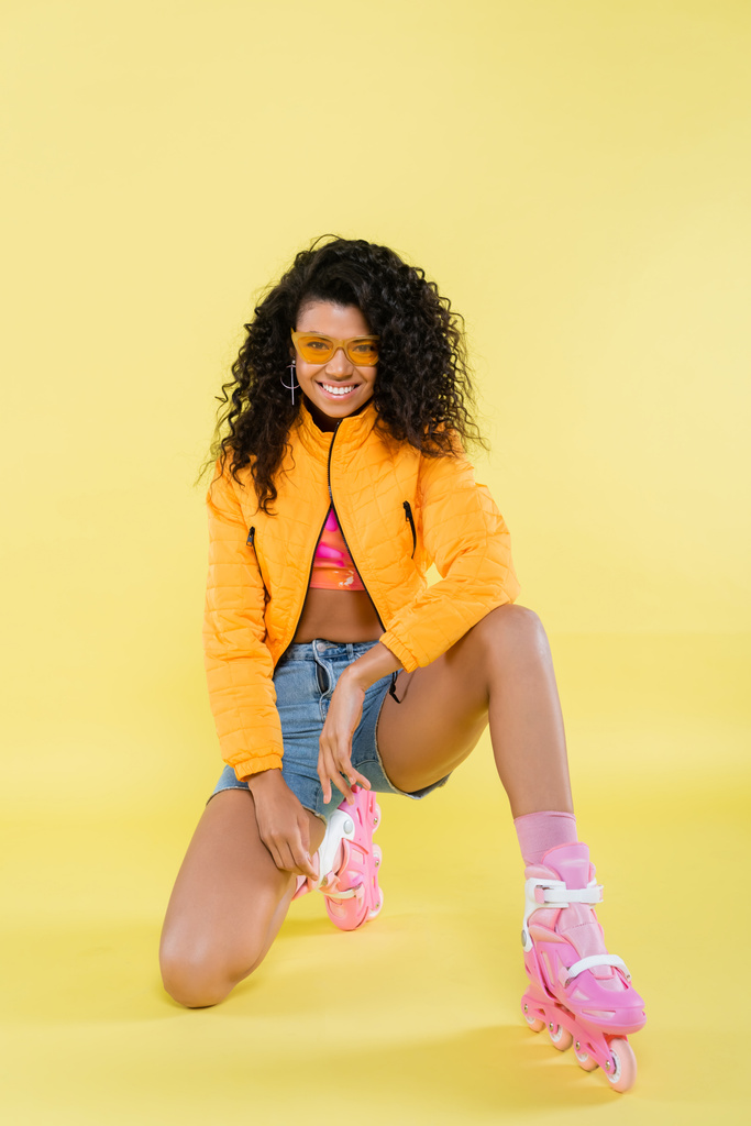 full length of smiling african american young woman in pink roller skates sitting on  knee on yellow - Photo, Image