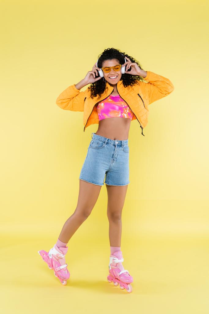 longitud completa de la joven afroamericana sonriente en patines de color rosa escuchando música en amarillo - Foto, imagen