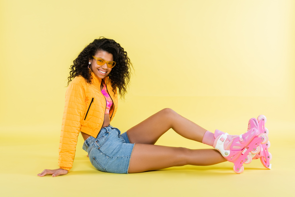 pleine longueur de heureuse jeune femme afro-américaine en lunettes de soleil et patins à roulettes roses assis sur jaune - Photo, image