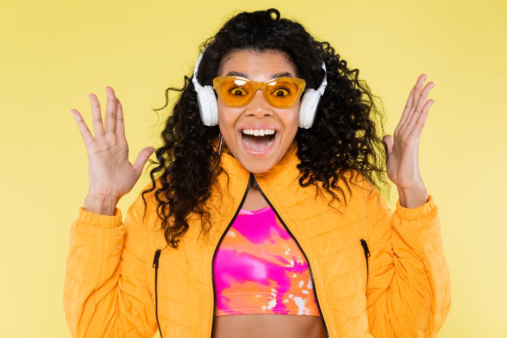 étonnante jeune femme afro-américaine en lunettes de soleil et écouteurs isolés sur jaune - Photo, image