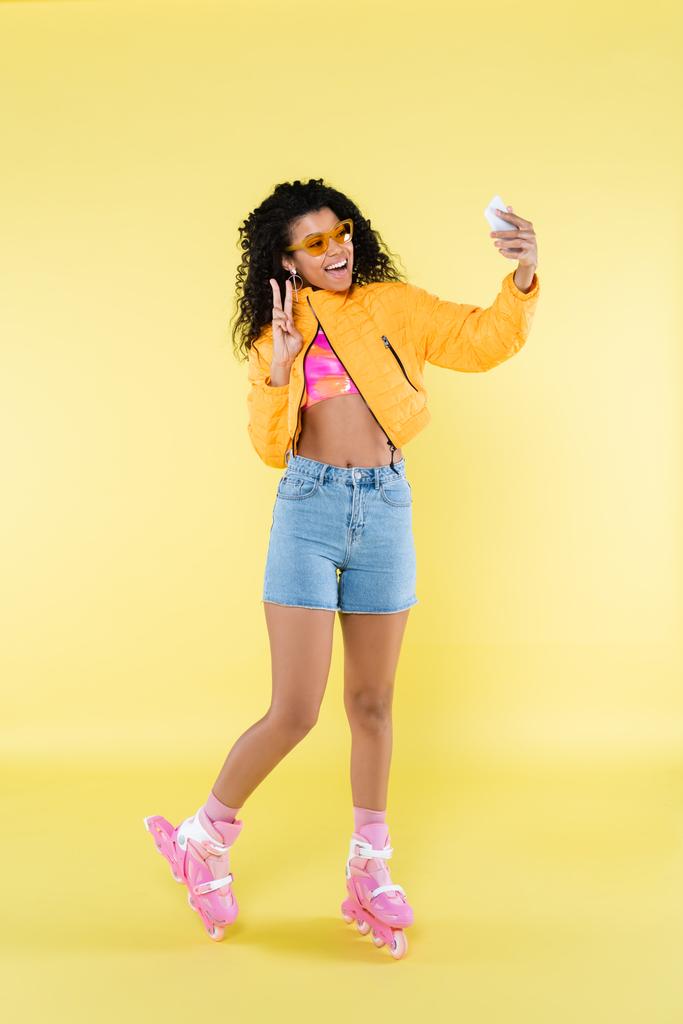 longitud completa de mujer joven afroamericana asombrada en patines de rodillo rosados que muestran el signo de la pieza mientras toma selfie en amarillo - Foto, imagen