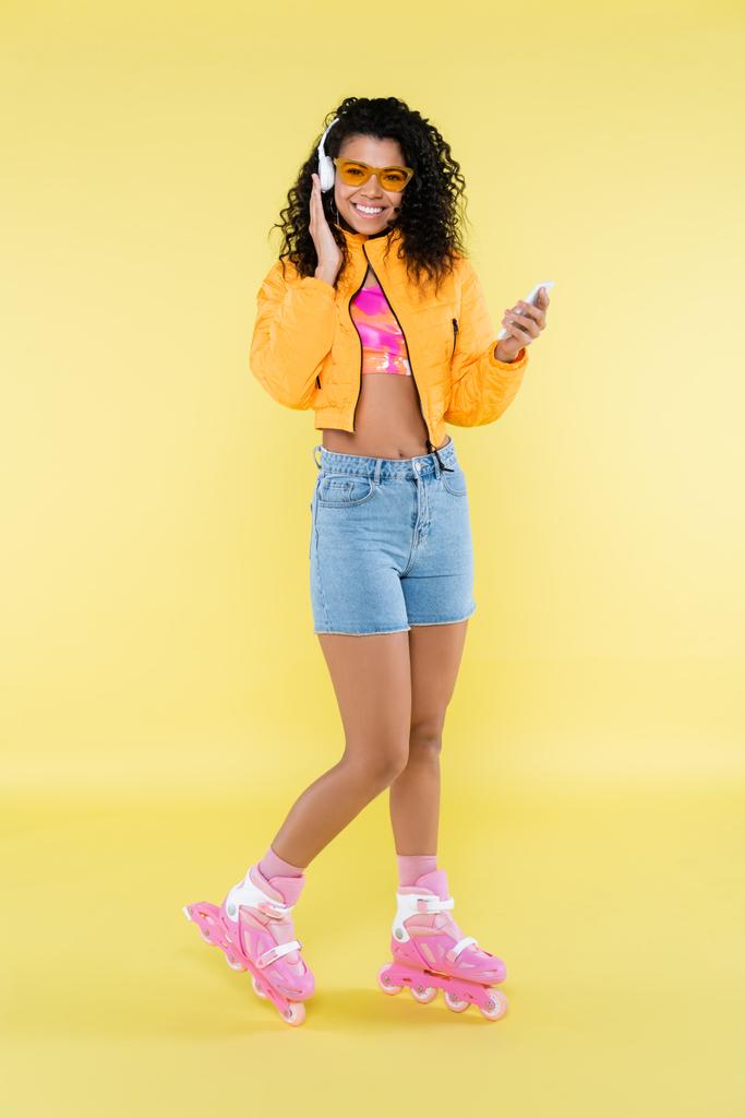 intera lunghezza della giovane donna afroamericana in pattini a rotelle rosa e cuffie utilizzando smartphone su giallo - Foto, immagini