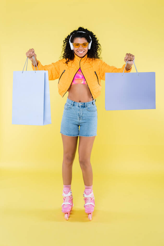 pełna długość afrykańskiej młodej kobiety w różowych wrotkach i słuchawkach z torbami na zakupy na żółto - Zdjęcie, obraz