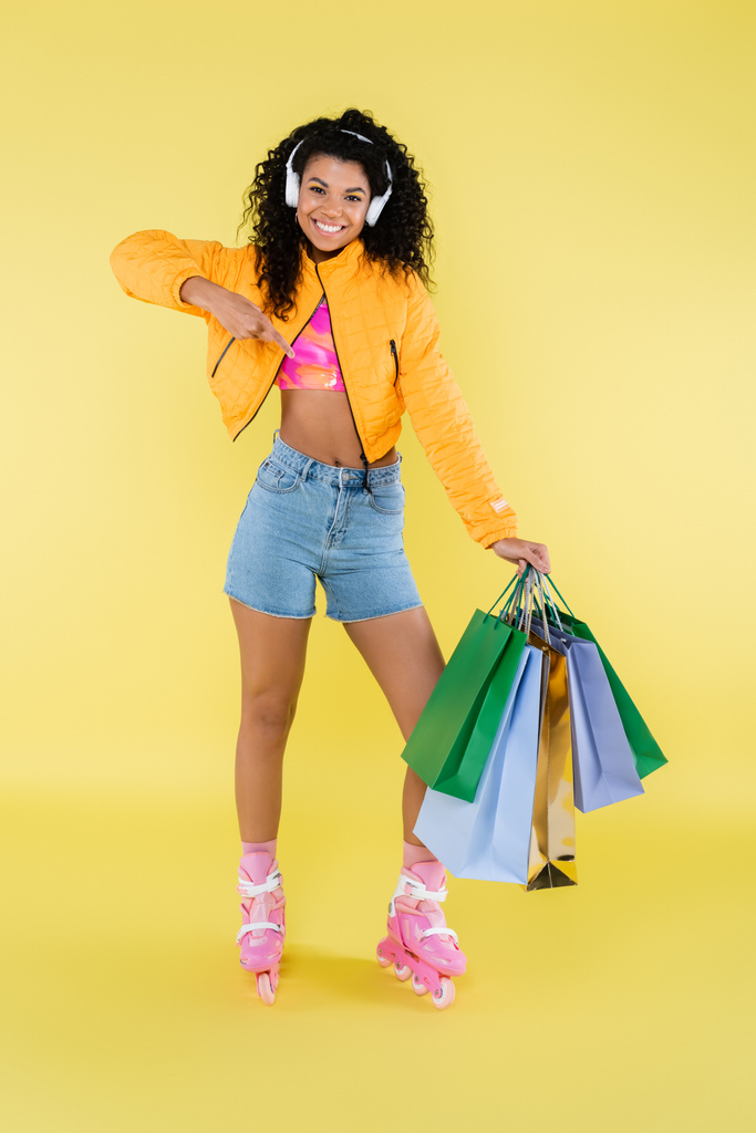 full length of happy african american young woman in roller skates and headphones pointing at shopping bags on yellow - Photo, Image