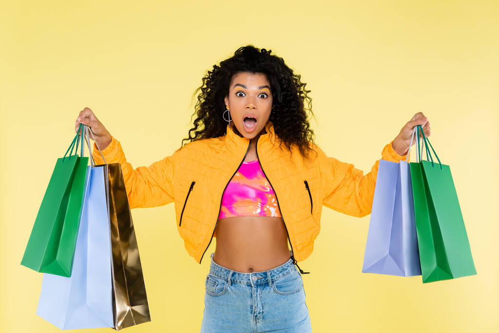 chocado afro-americano mulher segurando sacos de compras isolados no amarelo - Foto, Imagem