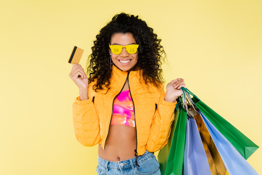 joyeuse jeune femme afro-américaine en lunettes de soleil avec vente lettrage tenant sacs à provisions et carte de crédit isolé sur jaune - Photo, image