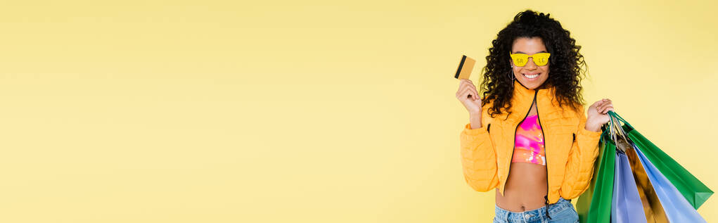 mujer afroamericana alegre en gafas de sol con letras de la venta sosteniendo bolsas de compras y tarjeta de crédito aislada en amarillo, bandera - Foto, imagen