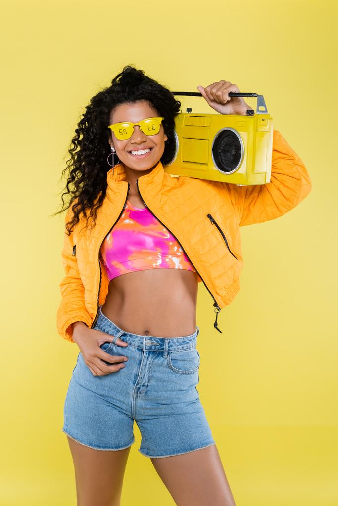 glimlachende Afrikaanse Amerikaanse jonge vrouw in een zonnebril met verkoop belettering bedrijf boombox geïsoleerd op geel - Foto, afbeelding