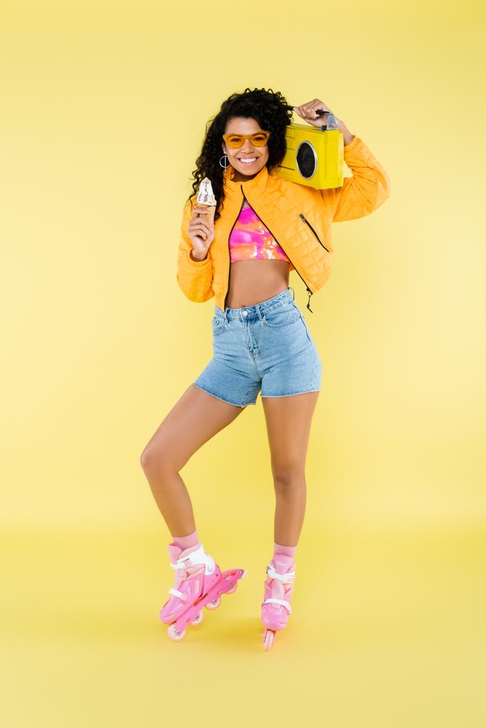 happy african american young woman in roller skates holding ice cream cone and boombox isolated on yellow - Photo, Image
