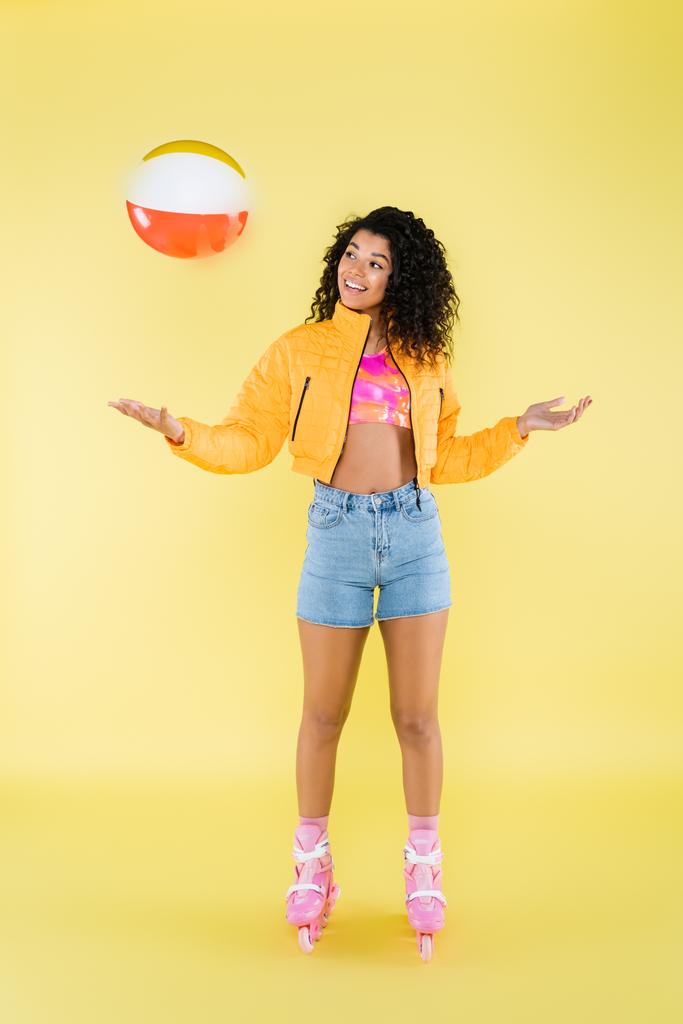 longitud completa de la joven africana americana positiva en patines lanzando la bola inflable en amarillo - Foto, Imagen