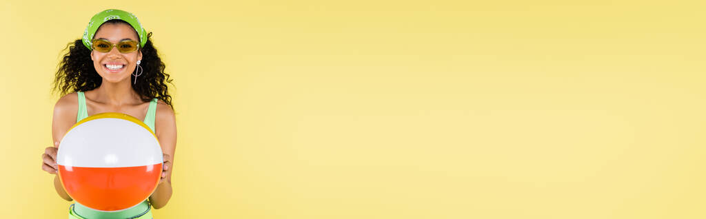 positive jeune femme afro-américaine en mouchoir et lunettes de soleil tenant ballon gonflable isolé sur jaune, bannière - Photo, image