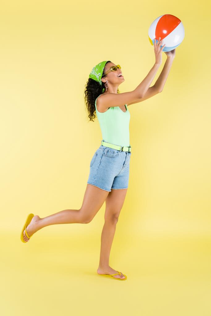 longitud completa de la mujer afroamericana feliz en gafas de sol que juegan con la bola inflable en amarillo - Foto, Imagen