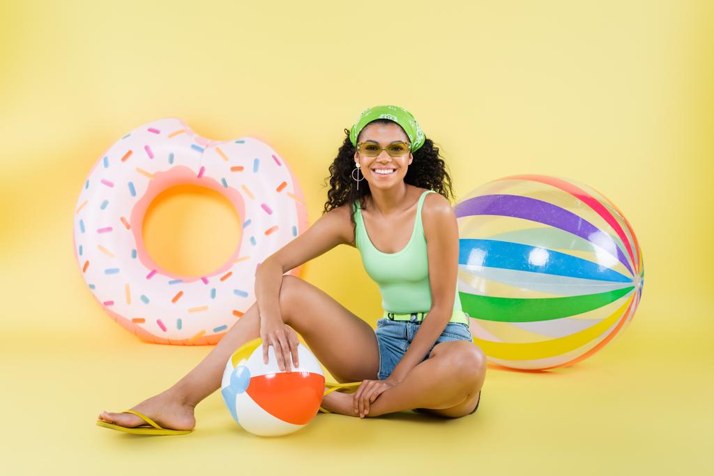 pełna długość szczęśliwy afrykański amerykański kobieta siedzi z plaża piłka na żółty - Zdjęcie, obraz