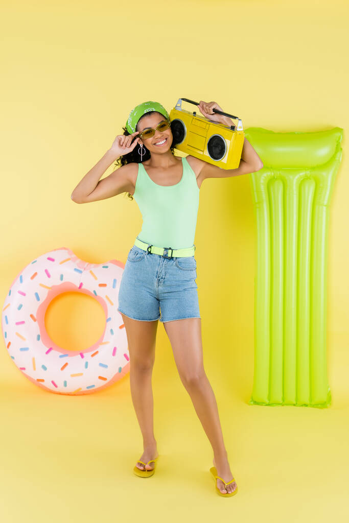longitud completa de la joven afro-americana alegre que sostiene el boombox cerca del colchón inflable y del anillo en amarillo  - Foto, Imagen