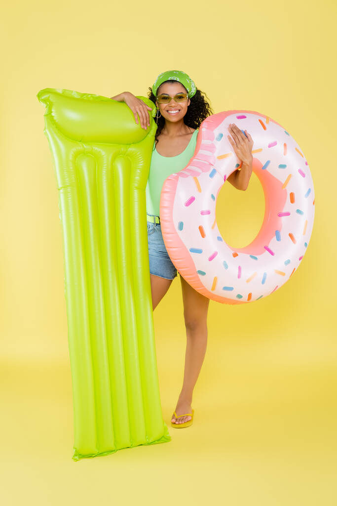 longitud completa de la mujer afroamericana feliz que sostiene el colchón inflable y el anillo en amarillo  - Foto, Imagen