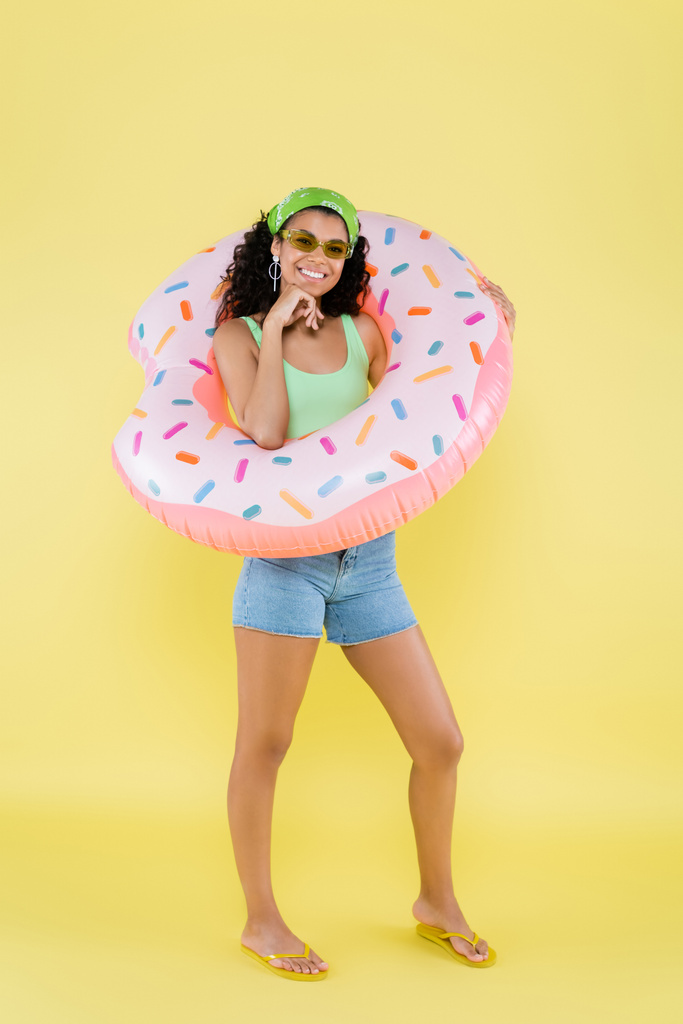 plná délka pozitivní afroamerické mladé ženy stojící s nafukovacím prstenem na žluté - Fotografie, Obrázek
