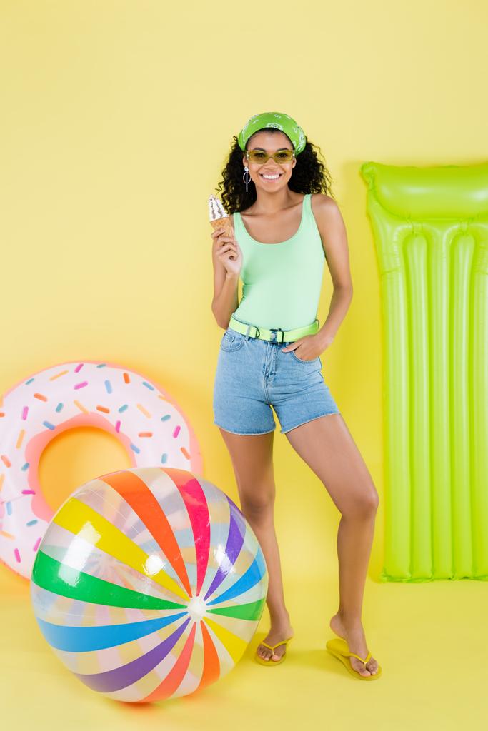 comprimento total da jovem americana africana positiva que está com cone de sorvete perto de bola inflável, colchão e anel no amarelo  - Foto, Imagem