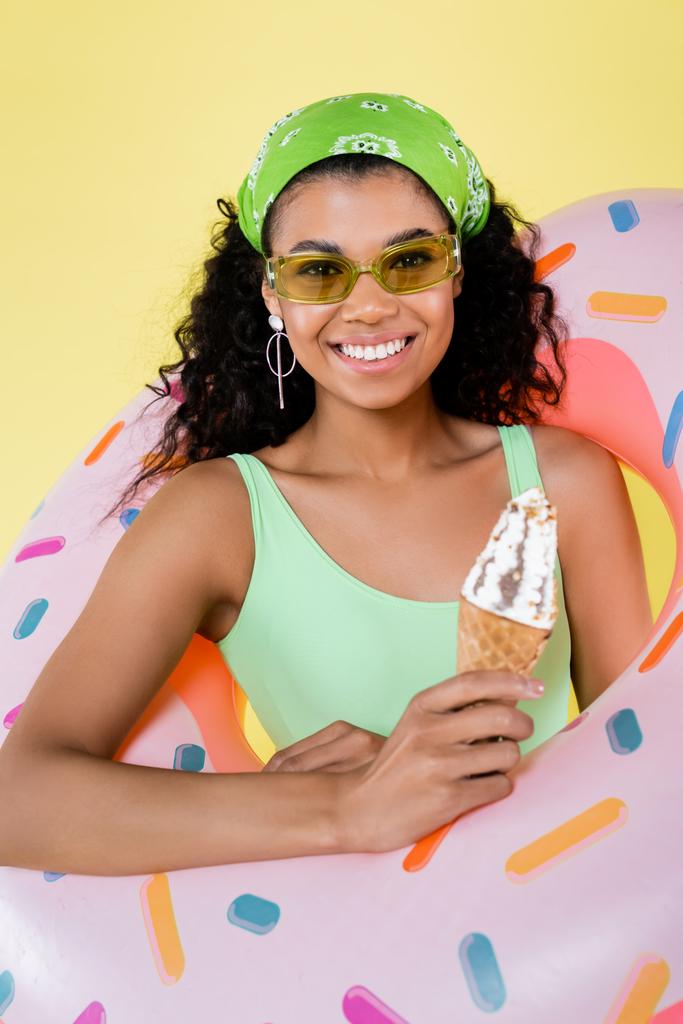 χαρούμενη αφροαμερικάνικη νεαρή γυναίκα στέκεται με φουσκωτό δαχτυλίδι και παγωτό χωνάκι απομονωμένο στο κίτρινο - Φωτογραφία, εικόνα