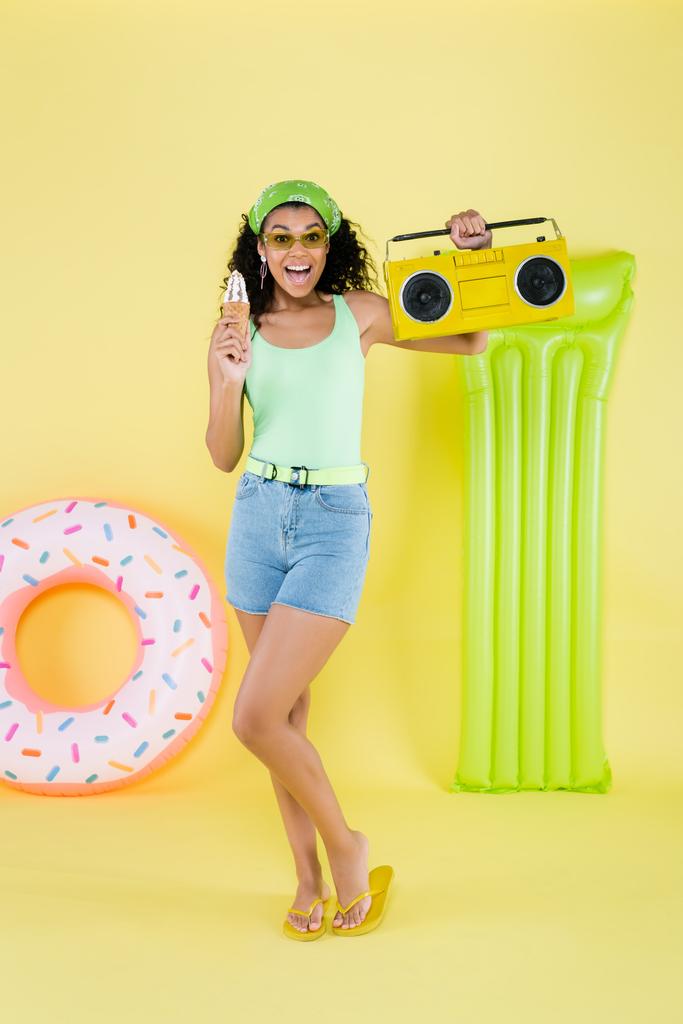 comprimento total da jovem americana africana alegre que está com boombox e cone de sorvete perto do anel inflável e do colchão no amarelo - Foto, Imagem