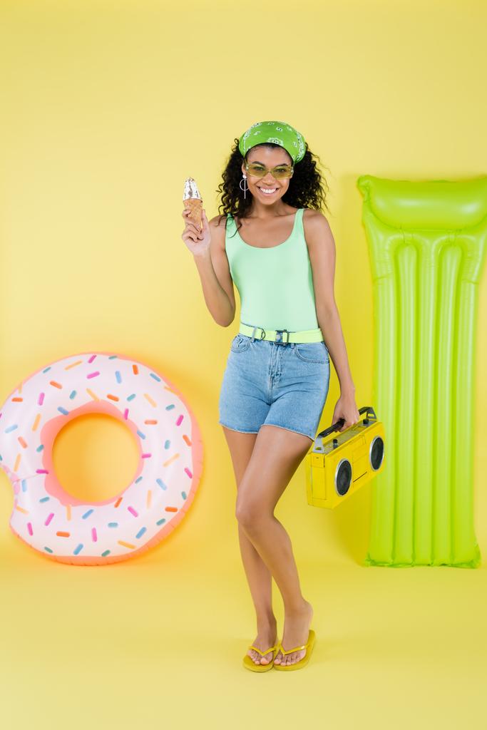 longitud completa de la joven afroamericana feliz que está parada con el boombox y el cono del helado cerca del anillo inflable y del colchón en amarillo - Foto, imagen