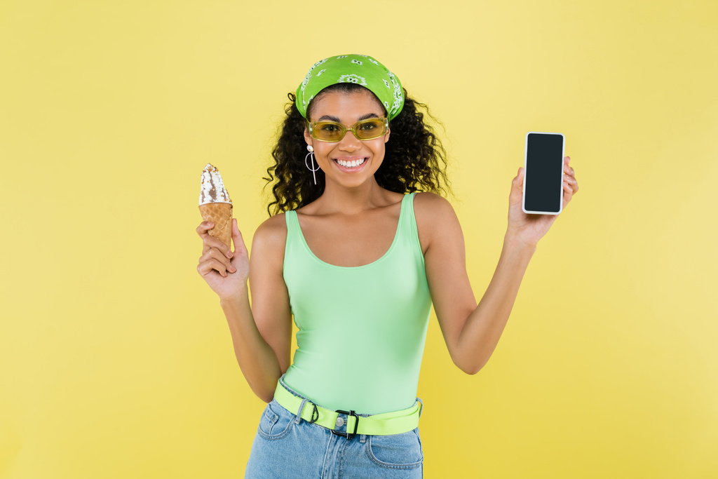 glückliche afrikanisch-amerikanische Frau mit Sonnenbrille und Eistüte und Smartphone mit leerem Bildschirm, isoliert auf gelb - Foto, Bild