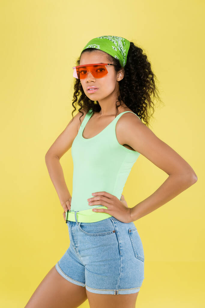 jong Afrikaans amerikaans jong vrouw in groen kerchief en zonnebril staan met de handen op heupen geïsoleerd op geel - Foto, afbeelding