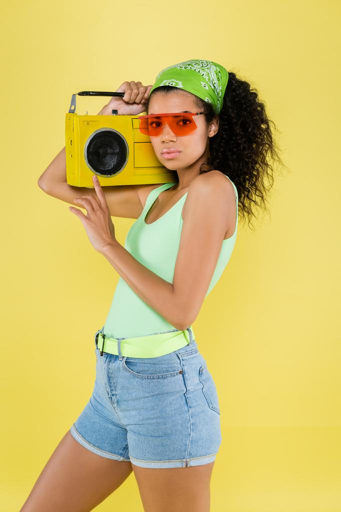 joven afroamericana joven en pañuelo verde y gafas de sol sosteniendo boombox aislado en amarillo - Foto, Imagen