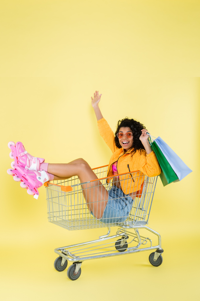 verbaasd Afrikaans amerikaanse jonge vrouw in roze rolschaatsen houden boodschappentassen terwijl zitten in winkelwagen op geel - Foto, afbeelding