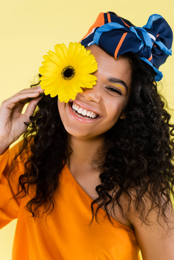 Χαρούμενη Αφροαμερικανή γυναίκα με μαντίλα που κρατά το λουλούδι απομονωμένο στο κίτρινο. - Φωτογραφία, εικόνα