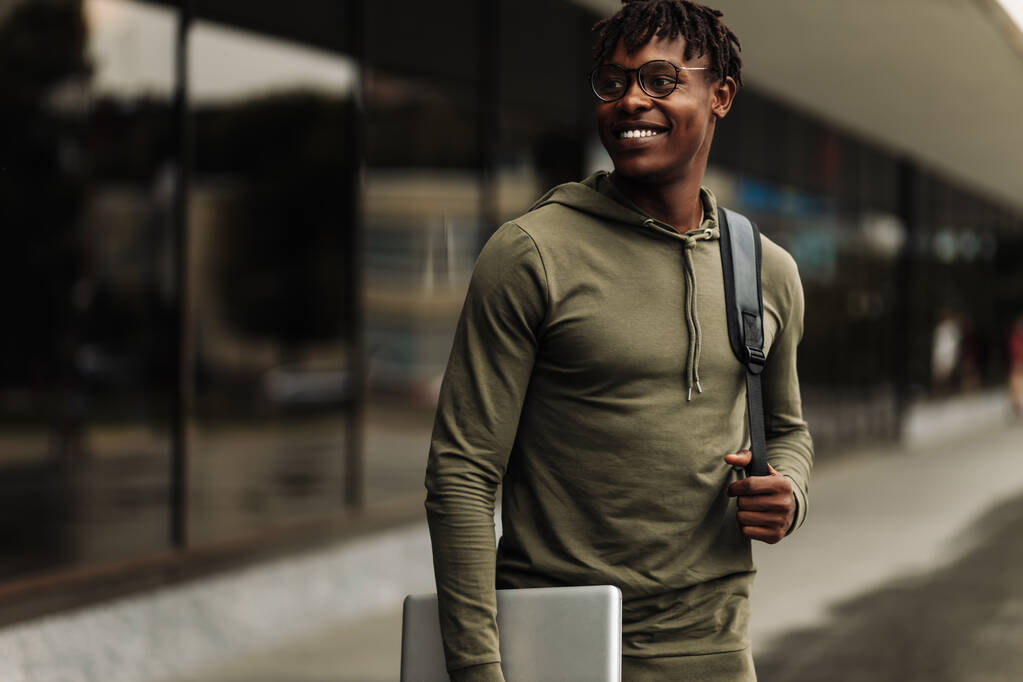 Guapo estudiante de piel oscura sonriente, con gafas con una mochila, sosteniendo una computadora portátil, caminando por el campus, descansando después de la escuela en el campus universitario - Foto, Imagen