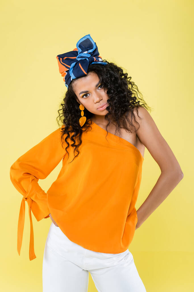curly african american woman in headscarf and bare shoulder posing with hands on hips isolated on yellow - Photo, Image