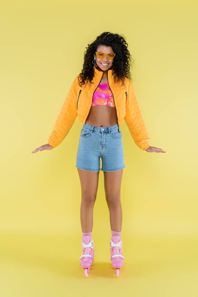 full length of positive and curly african american young woman in pink roller skates on yellow - Photo, Image
