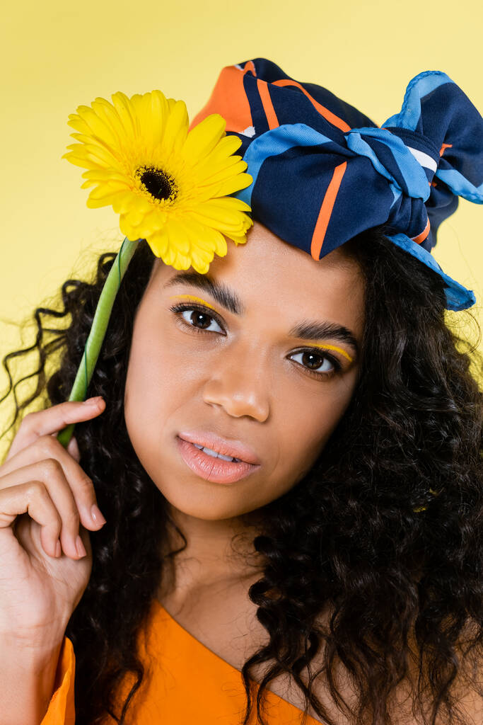кудрявая африканская американка в платке держит цветок изолирован на желтый - Фото, изображение