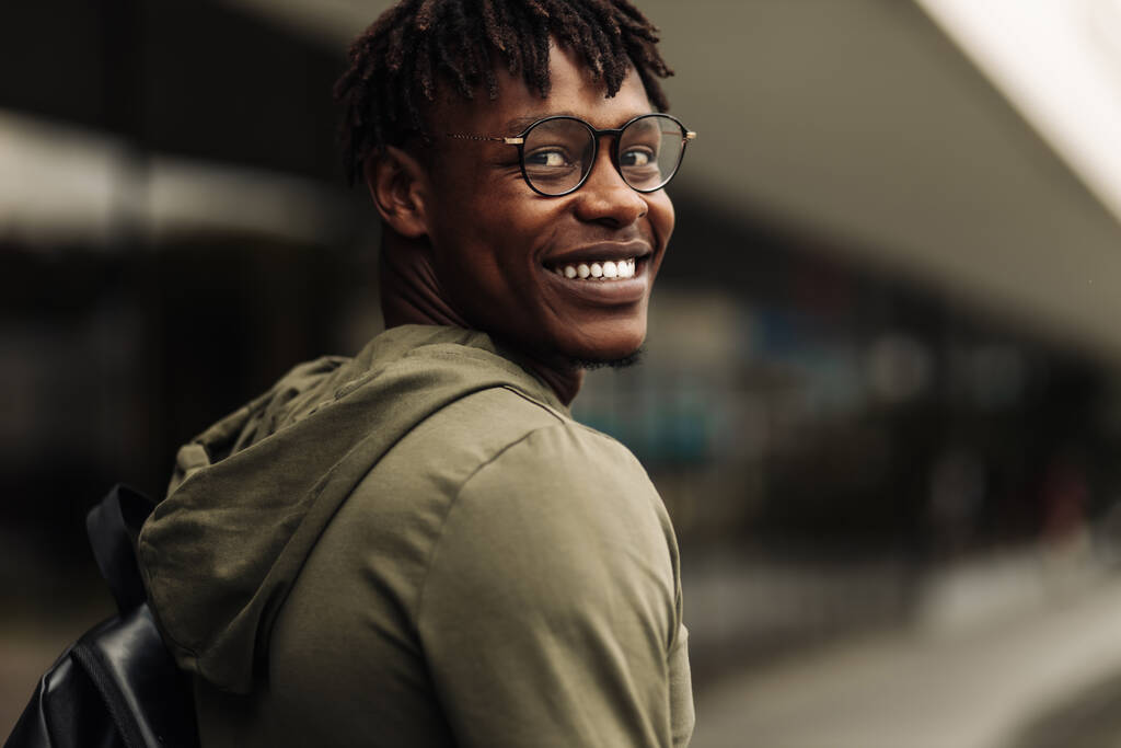 Gelukkige knappe succesvolle Afrikaanse student, het dragen van een bril en met een zwarte rugzak, staande op de trap en glimlachend, kijkt in het frame buiten op straat in de buurt van de universiteit - Foto, afbeelding