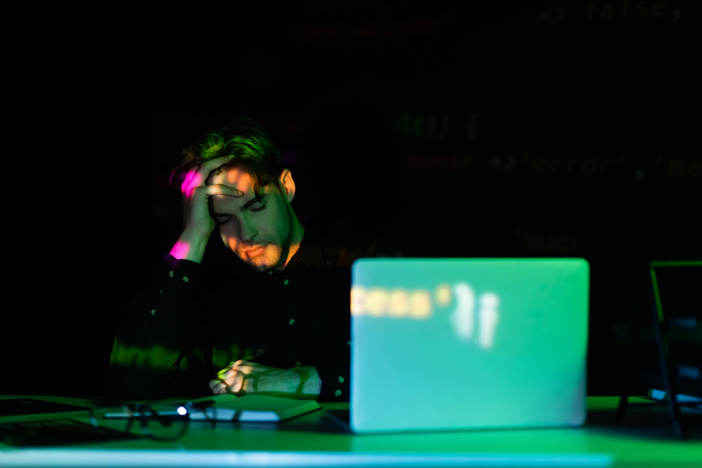 Programador sentado cerca de portátil y portátil con reflejo de luz aislado en negro  - Foto, imagen