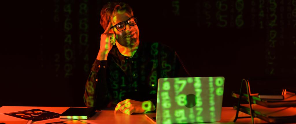 Usmívající se programátor v brýlích při pohledu na notebook s odrazem počítačového kódu v blízkosti papíry na černém pozadí, banner  - Fotografie, Obrázek