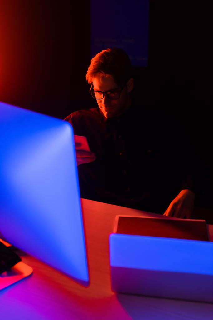 Programmierer in Brille mit Smartphone in der Nähe von Computern auf Tisch auf schwarzem Hintergrund  - Foto, Bild