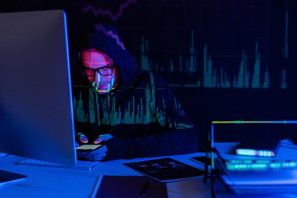 Hacker con reflejo de gráficos usando smartphone cerca del ordenador y documentos sobre fondo negro  - Foto, Imagen
