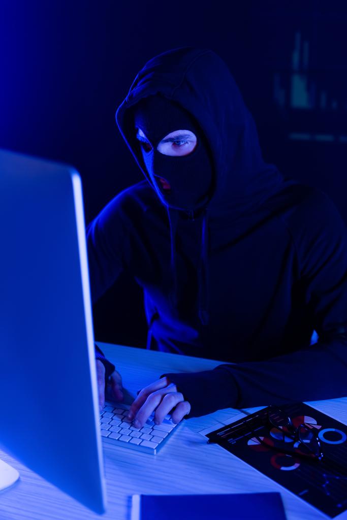 Hacker en pasamontañas usando computadora cerca de papeles en la mesa sobre fondo negro  - Foto, imagen