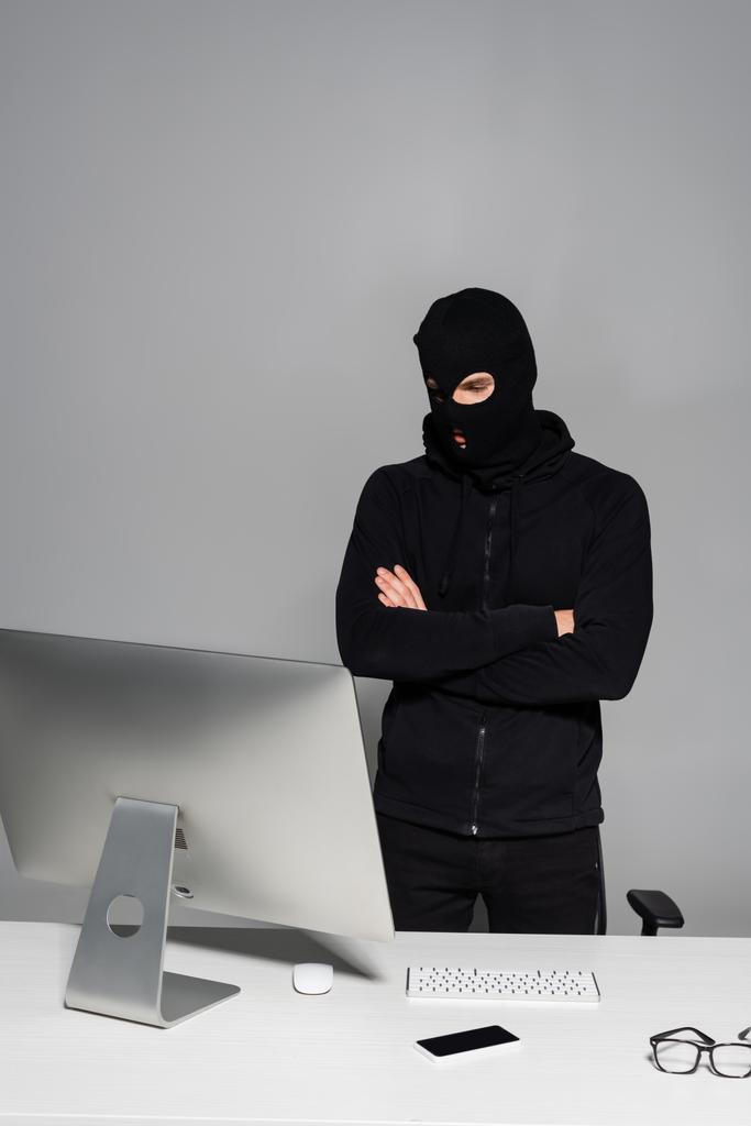 Hacker en pasamontañas de pie con los brazos cruzados cerca de la computadora y el teléfono inteligente en la mesa aislado en gris  - Foto, imagen
