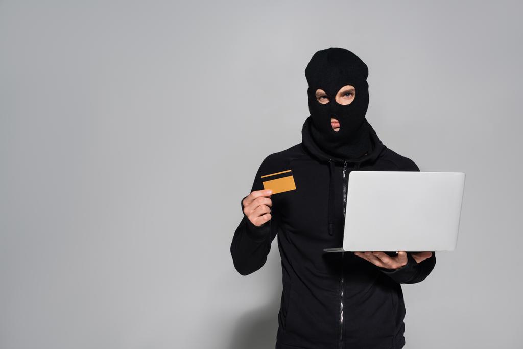 バラクラバのハッカーはクレジットカードとラップトップを持っています灰色の背景  - 写真・画像
