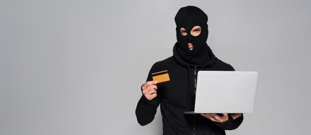 Hacker in balaclava holding laptop and credit card isolated on grey, banner  - Photo, Image