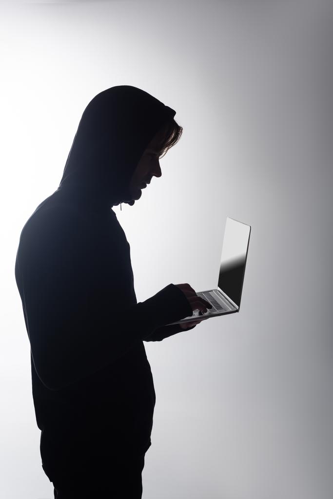 Vista laterale di hacker in cappuccio utilizzando laptop isolato su grigio  - Foto, immagini