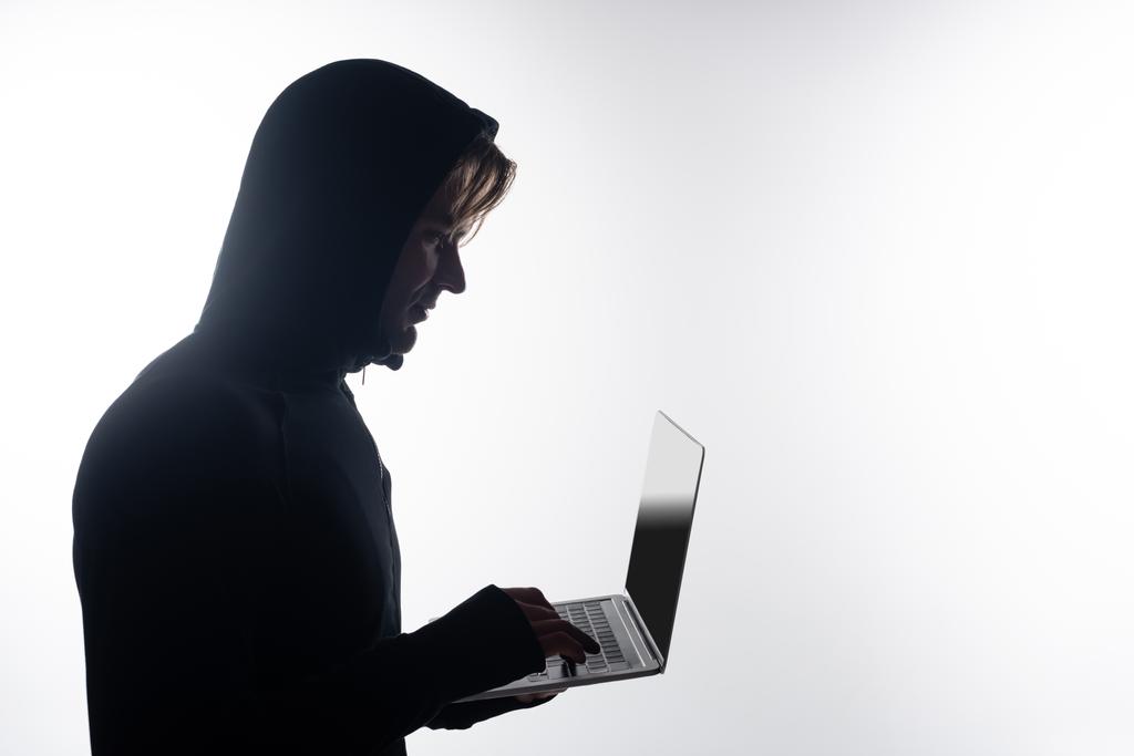 Boční pohled hackera v kapuci pomocí notebooku s prázdnou obrazovkou izolovanou na šedé  - Fotografie, Obrázek