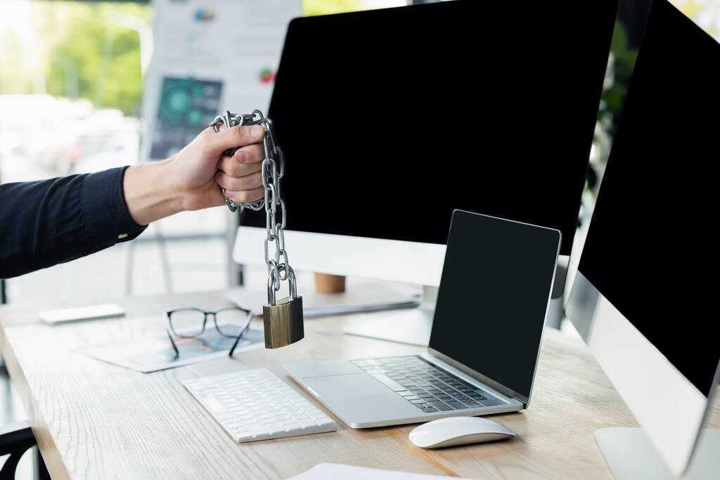 Cropped view of programmer holding padlock on chain near computers with blank screen in office  - Photo, Image