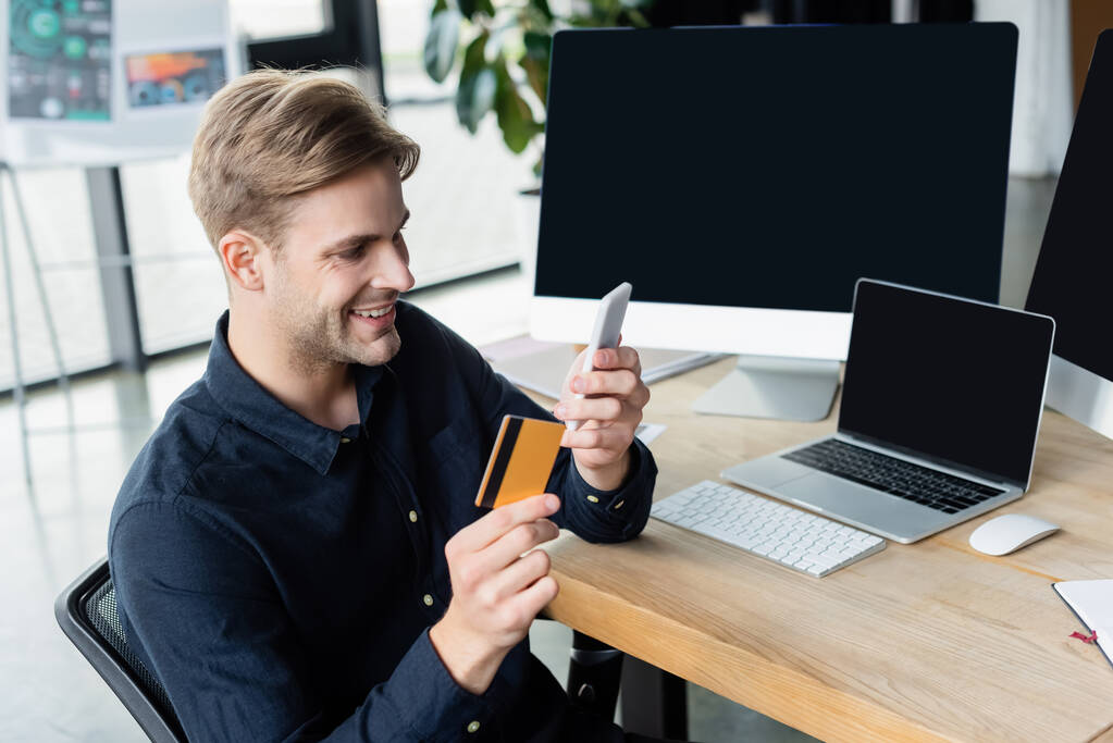Позитивный разработчик, использующий смартфон и кредитную карту рядом с компьютерами в офисе  - Фото, изображение