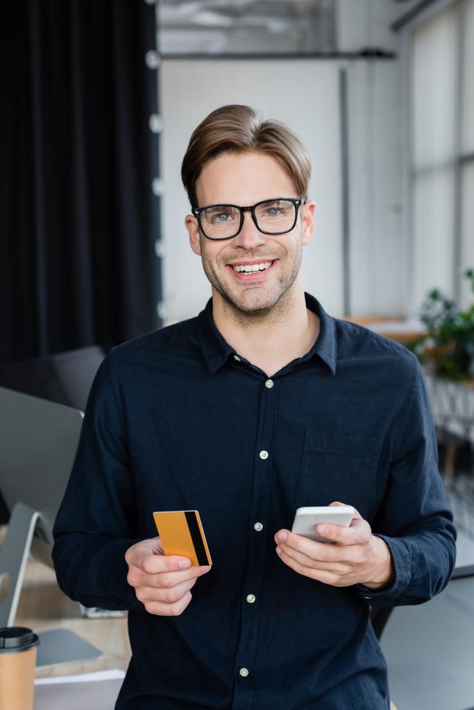 Glimlachende ontwikkelaar met creditcard en mobiele telefoon terwijl hij naar de camera op kantoor kijkt  - Foto, afbeelding