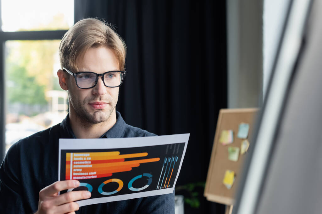 Young programmer holding document near blurred flip chart in office  - Photo, Image